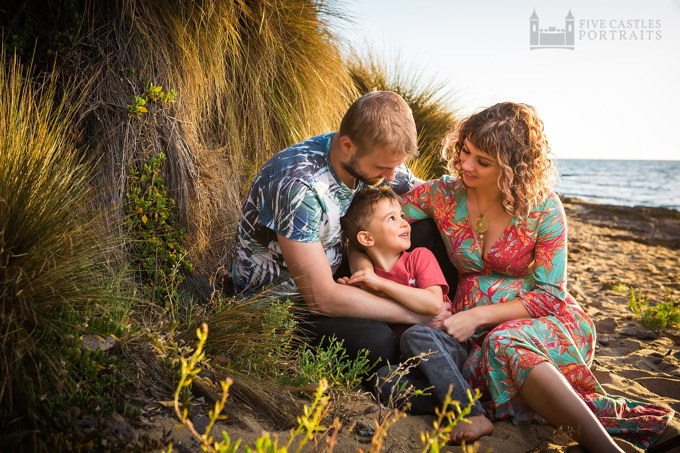 A family of three at golden hour on the beach, the father kissing his son's head while the expectant mother looks on, at a Five Castles Portraits session