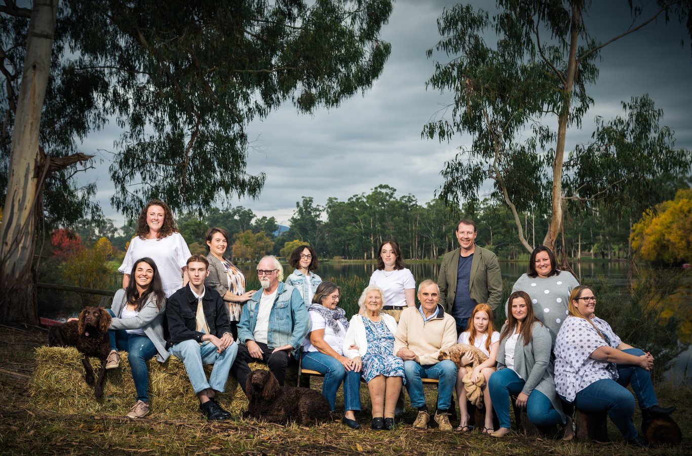 Large multi-generational family (14+ people) with two dogs seated by water with eucalyptus trees
