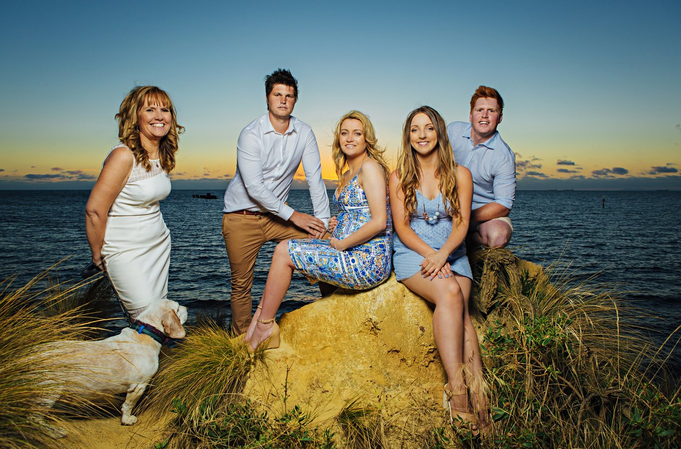 Large family group (five people plus dog) naturally arranged on rocks at golden hour