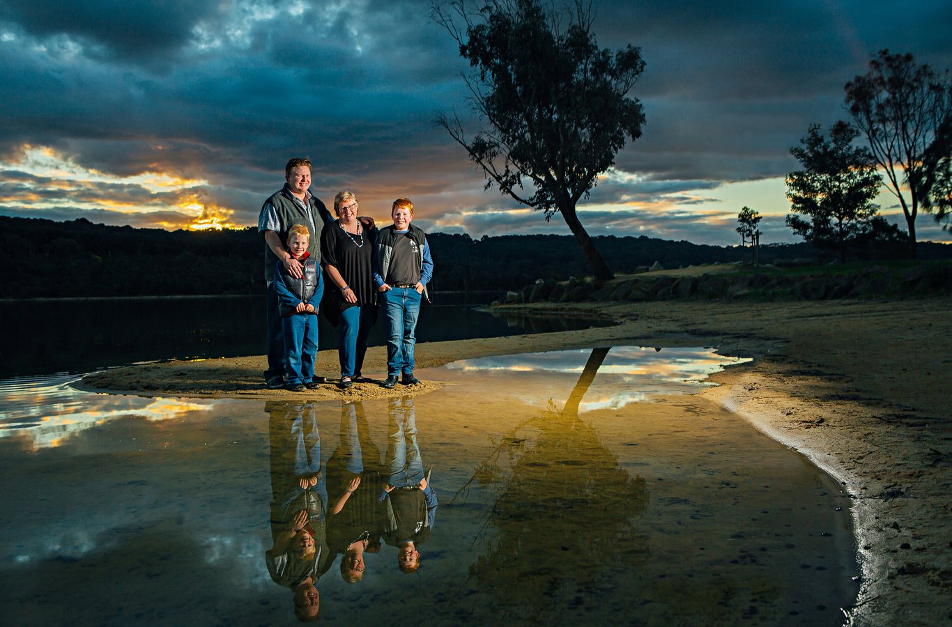 Family of four by river at golden hour with dramatic sky and water reflections; strong environmental composition